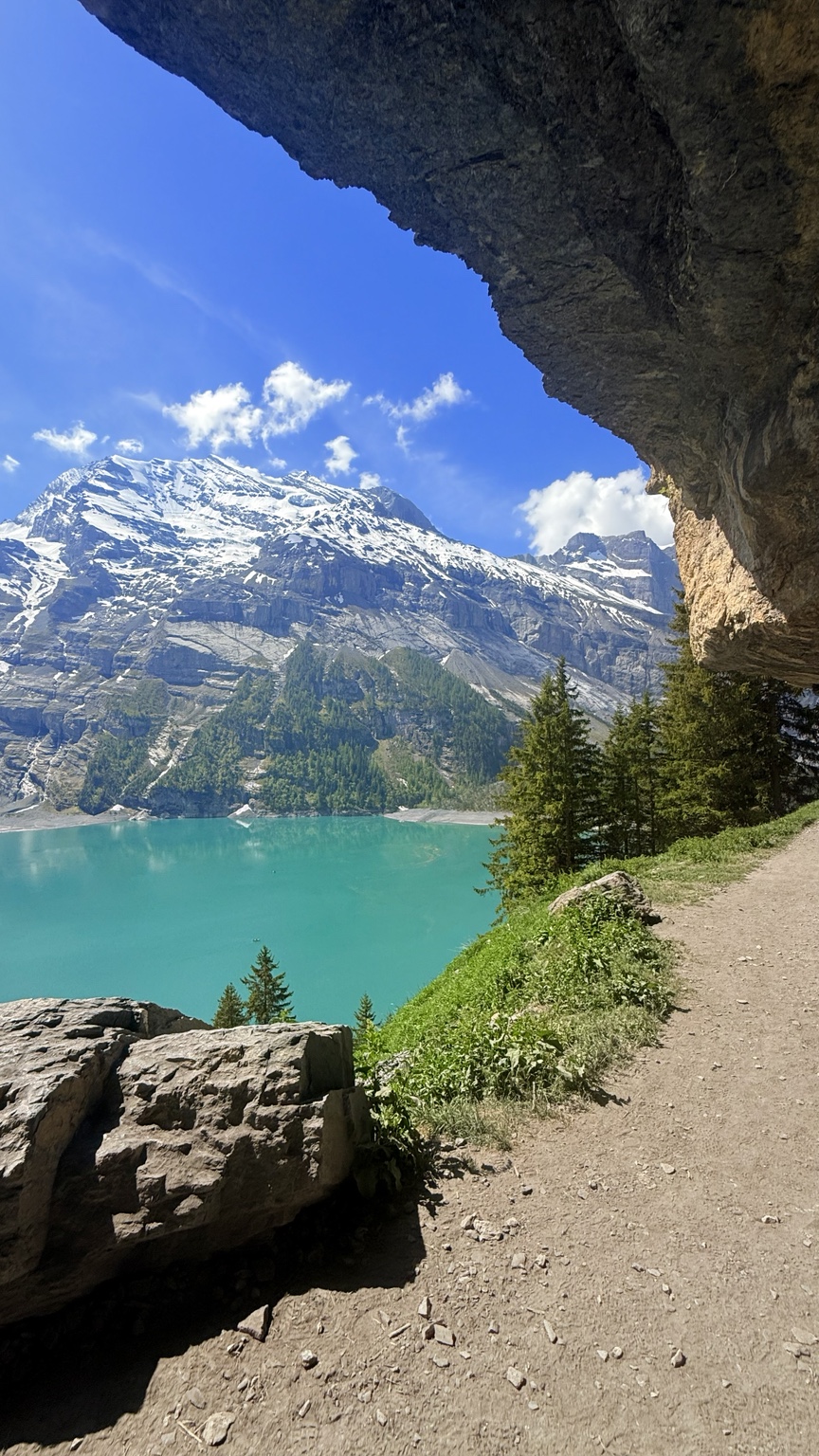 Trail beneath a rock overhang looking across the turquoise water of Oeschinensee