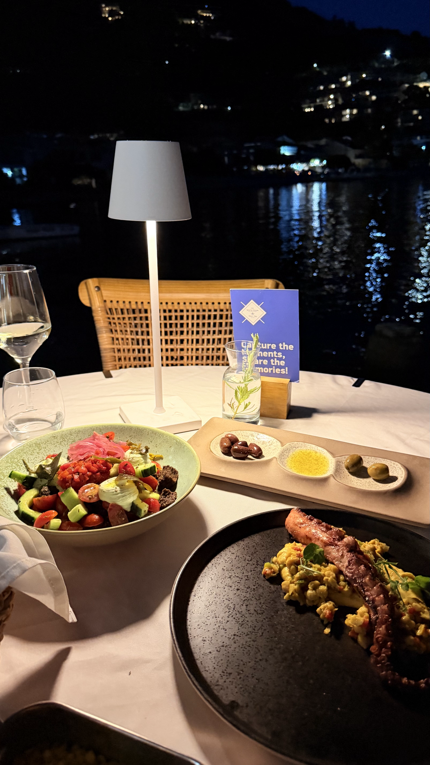 An evening table with a Greek salad, olive tray and octopus dish, the bay dark behind