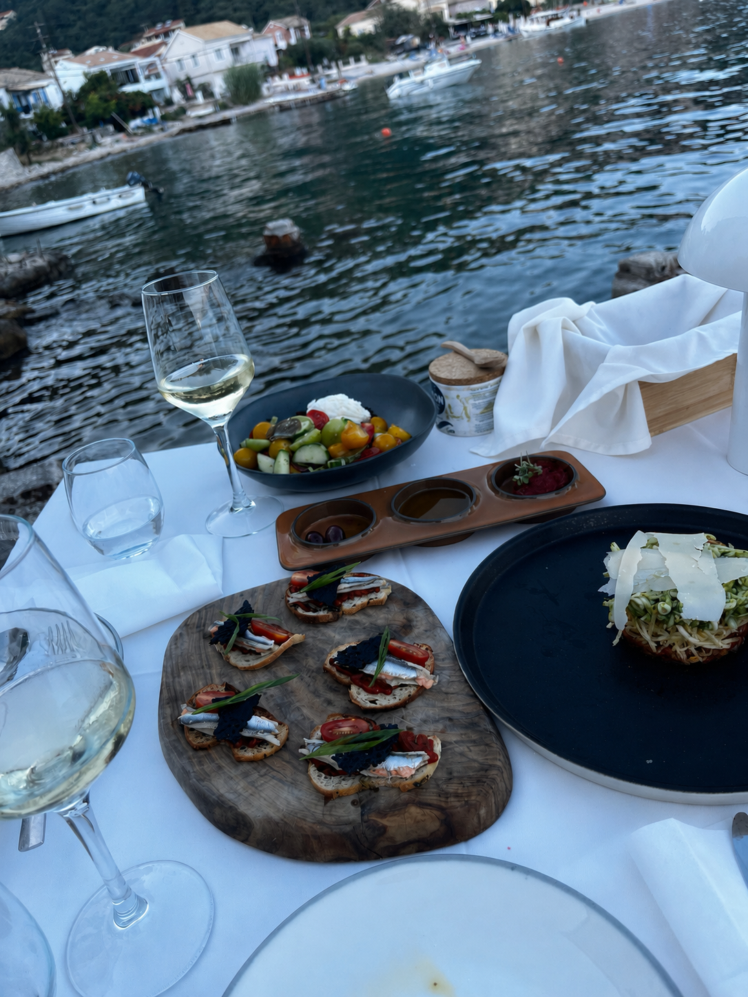 A daytime table on the water with a Greek salad, olive oil tasting tray and starters, Kalami Bay behind